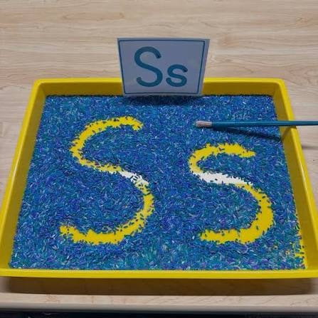 The letter S traced twice in a tray of colored rice, with an Ss letter card behind it