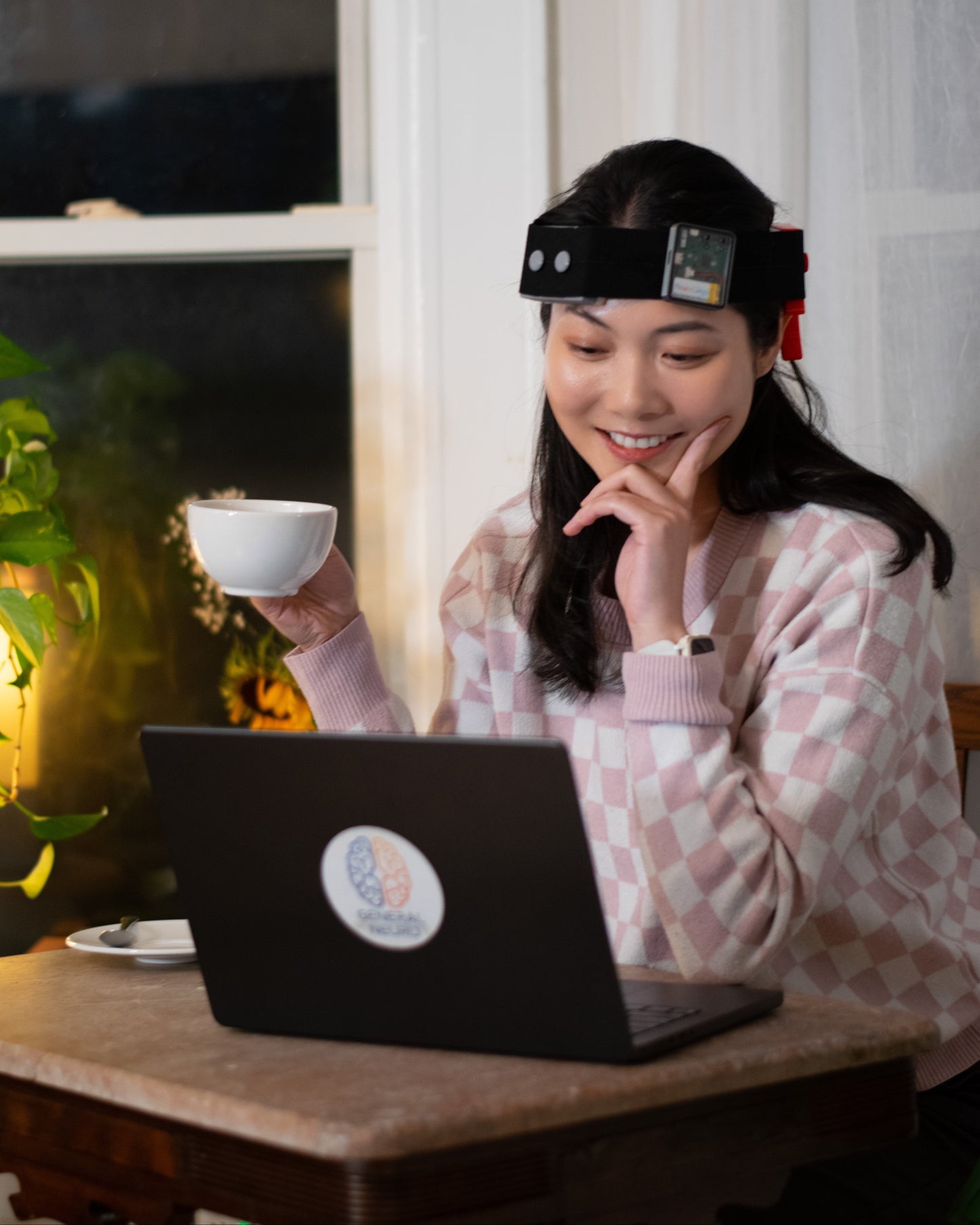 Woman sitting at a table with a laptop, holding a cup, and wearing a brain stimulation headset, the NeuroLingo Model 1. 