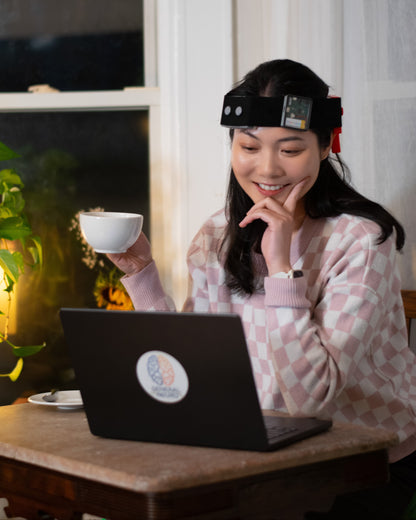 Woman sitting at a table with a laptop, holding a cup, and wearing a brain stimulation headset, the NeuroLingo Model 1. 