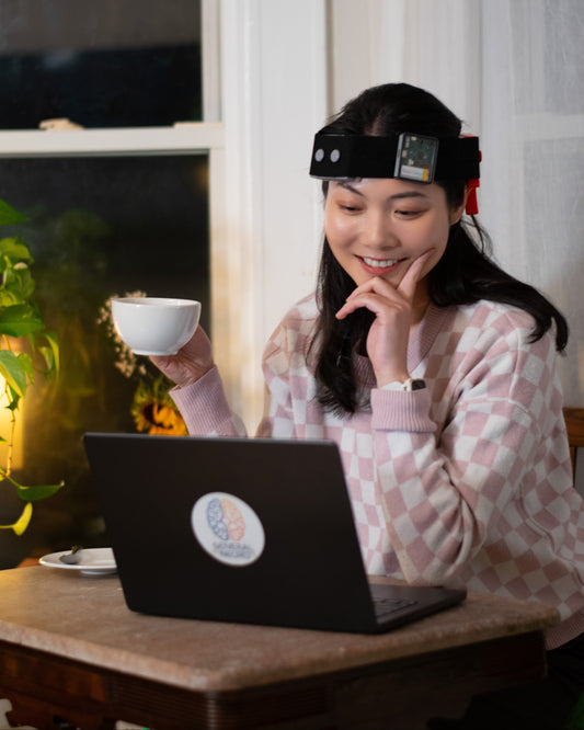 Woman sitting at a table with a laptop, holding a cup, and wearing a brain stimulation headset, the NeuroLingo Model 1. 