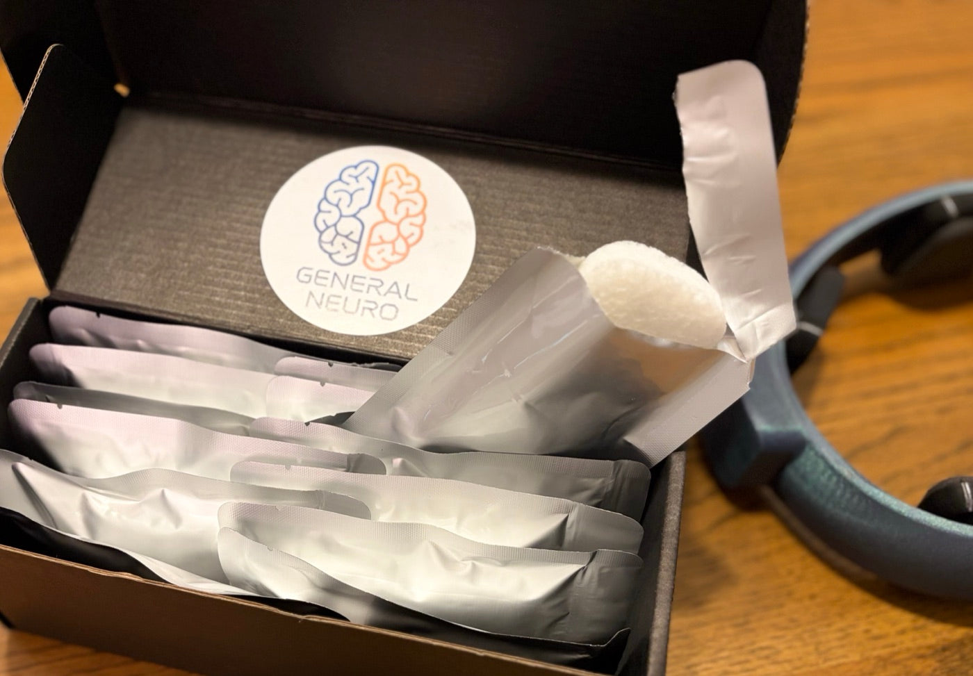 A NeuroLingo refill box of sealed foil session packs, one opened to show the white sponge pad, next to the headband